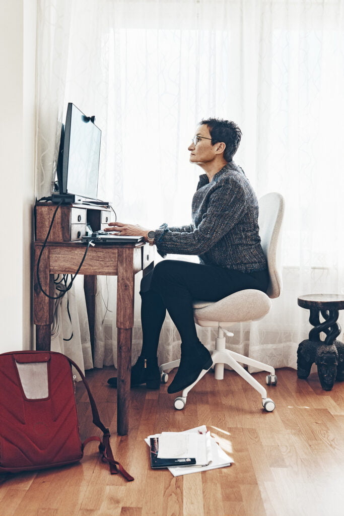 En kvinna sitter vid ett litet skrivbord i hennes hemmakontor. Vita gardiner i bakgrunden.